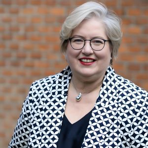 Lächelnde Frau mit kurzem, hellgrauem Haar und runder Brille, die ein schwarzes Oberteil und einen weißen Blazer mit einem geometrischen schwarzen Muster trägt, steht vor einem unscharfen Backsteinmauer-Hintergrund.